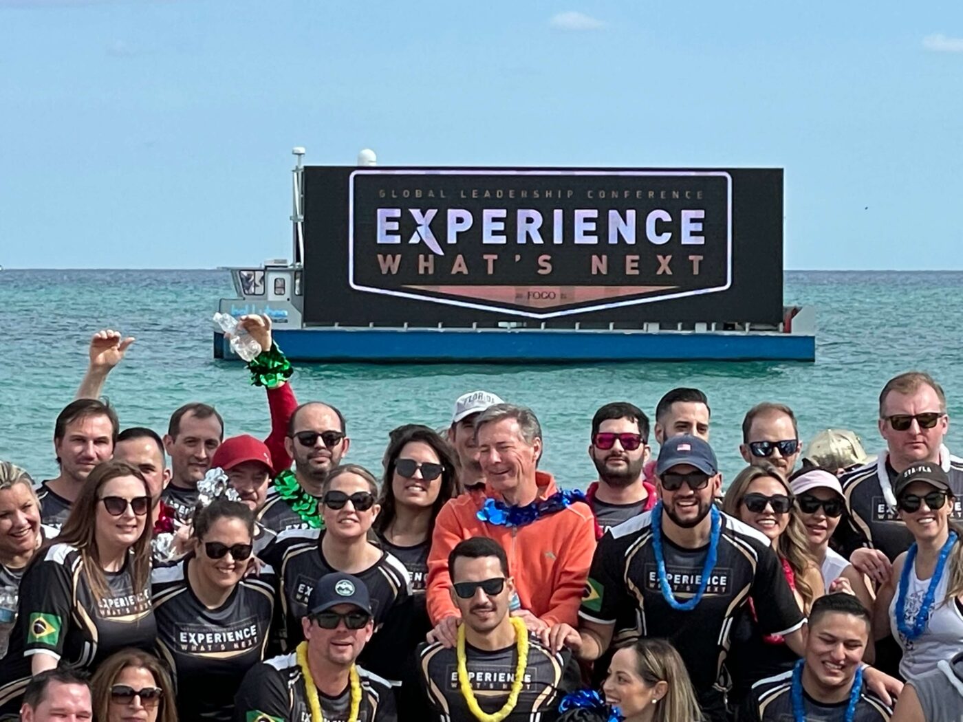 Group in black shirts on beach posing by boat with Global Leadership Conference sign; outdoor advertising for Corporate Events.