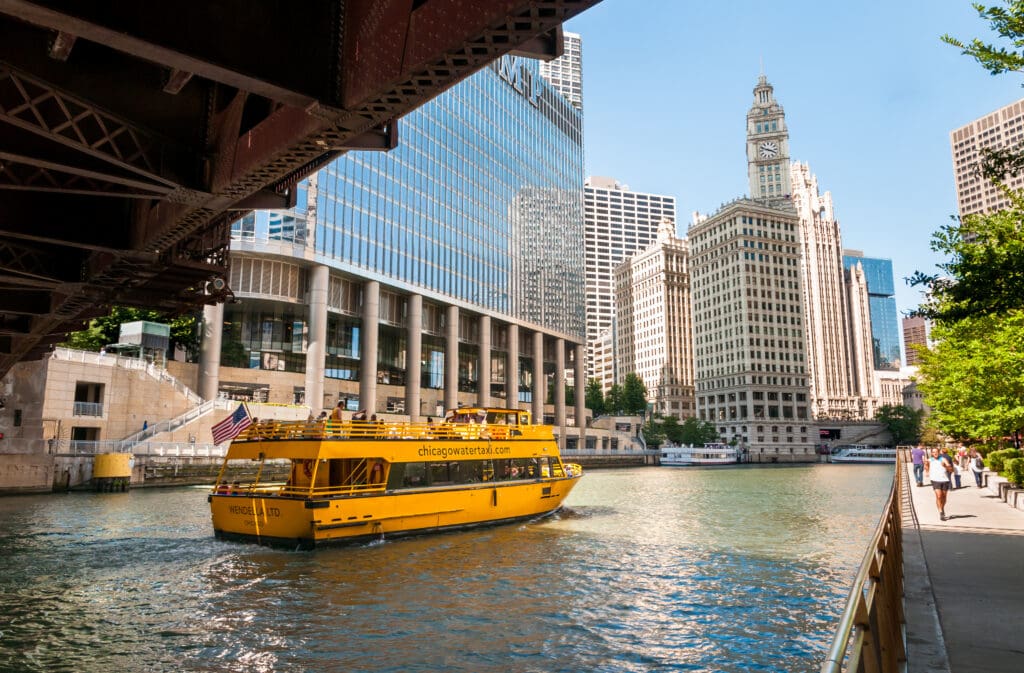Chicago Water Taxi sailing along the Riverwalk adjacent to high-rise offices and outdoor seating near the Wacker Drive corridor, a key zone for targeting business professionals.
