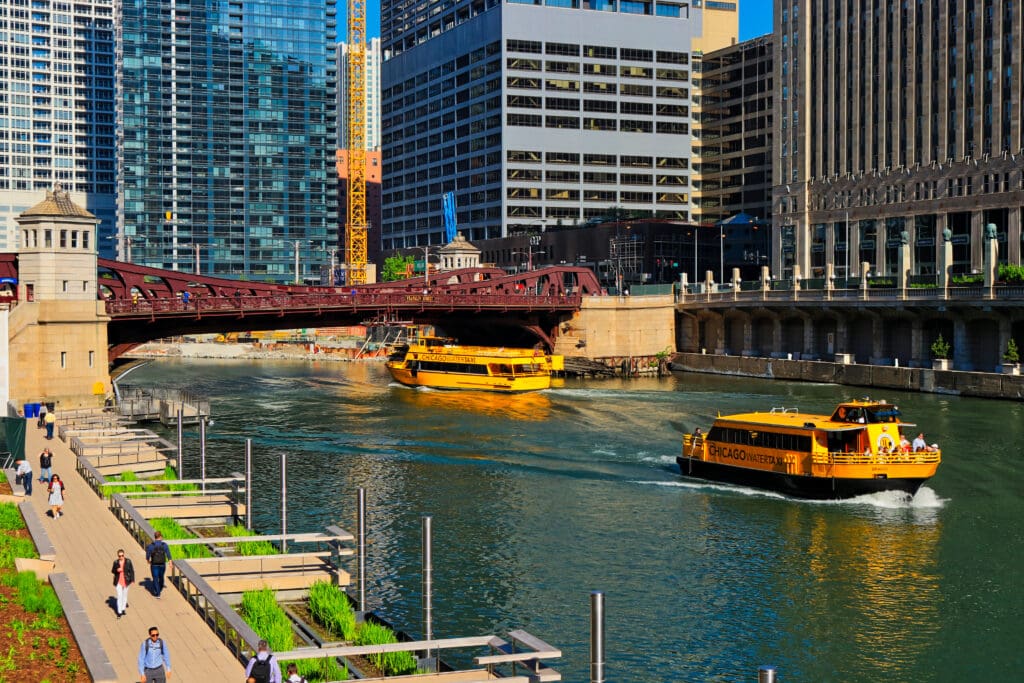 Chicago Water Taxi cruising past the Chicago Riverwalk and Clark Street Bridge, a high-traffic pedestrian and leisure zone ideal for downtown OOH advertising visibility.