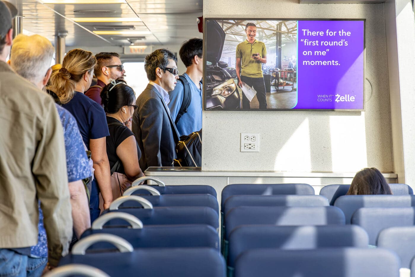 Passengers stand inside a ferry near blue seats; DOOH transit advertising shows a Zelle ad with "first rounds on me" message.