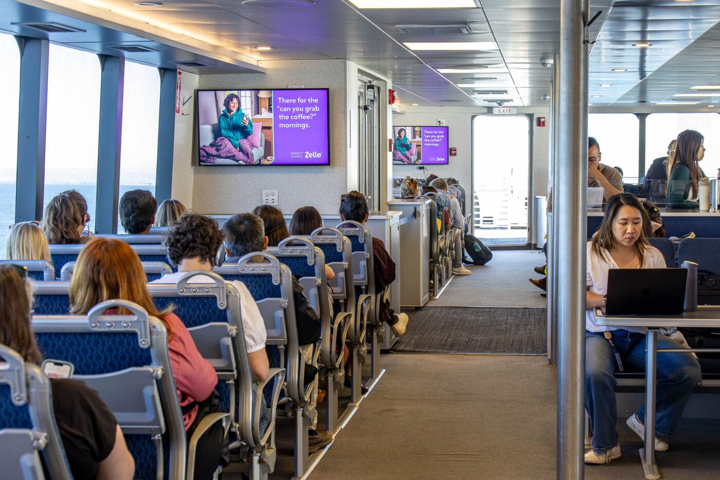 Passengers in blue seats on a ferry see DOOH transit advertising screens showing a woman on a sofa; large windows run along the left.