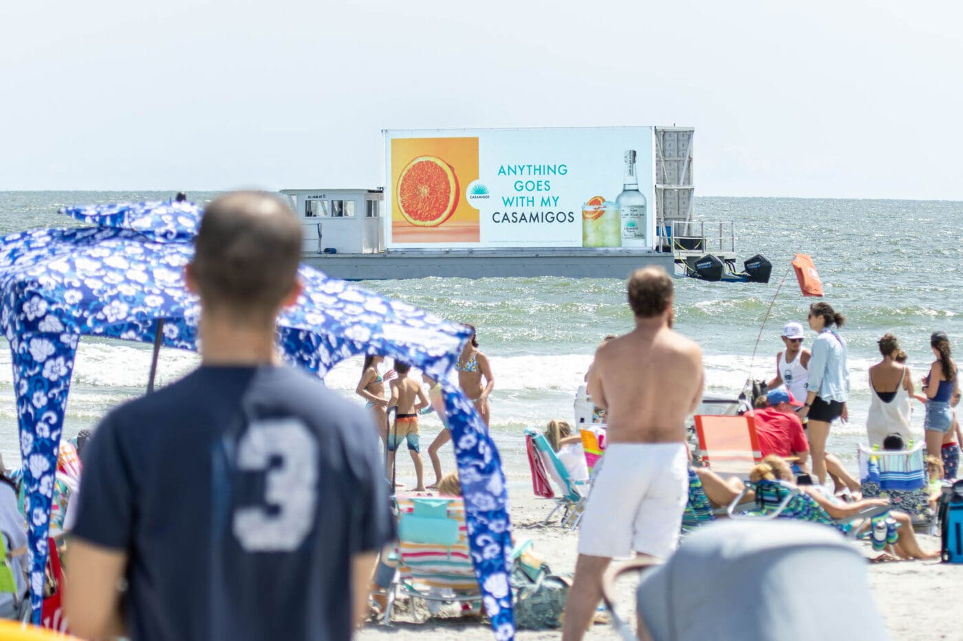 Beachgoers watch a crowded shore as a floating Casamigos tequila transit advertising billboard passes by on the ocean, promoting the brand.