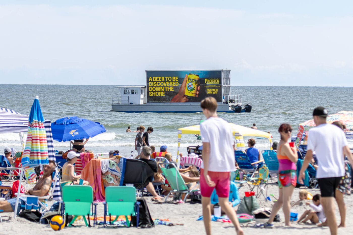 Crowded beach with colorful chairs, umbrellas, and a ferry featuring outdoor billboard advertising for beer on the ocean near swimmers.