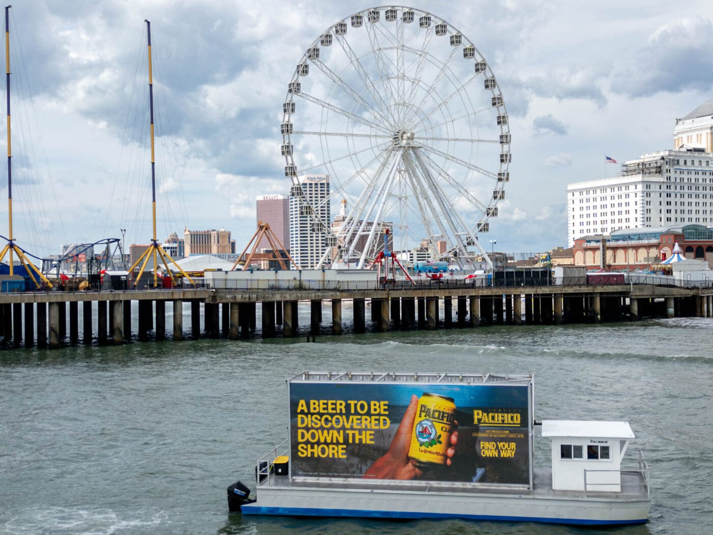 Ferry with Pacifico beer transit advertising sails near a pier, Ferris wheel, and buildings under cloudy skies—outdoor OOH ad exposure.