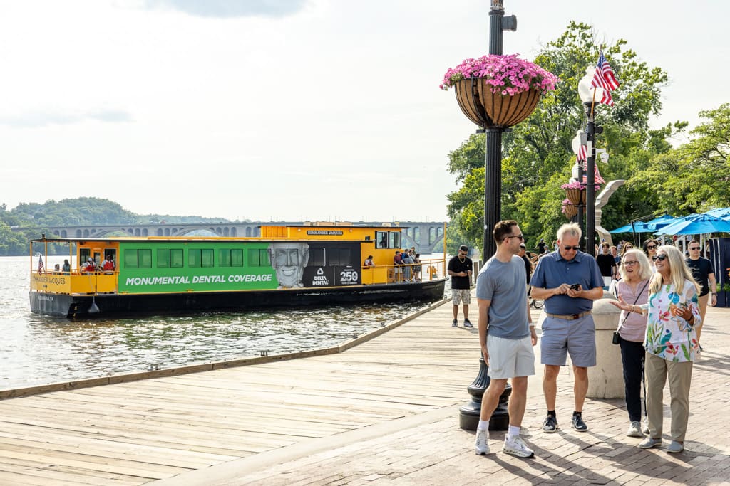 Four people walk on a wooden riverside path by a docked boat featuring transit advertising for dental coverage; OOH signage, flags, flower baskets nearby.